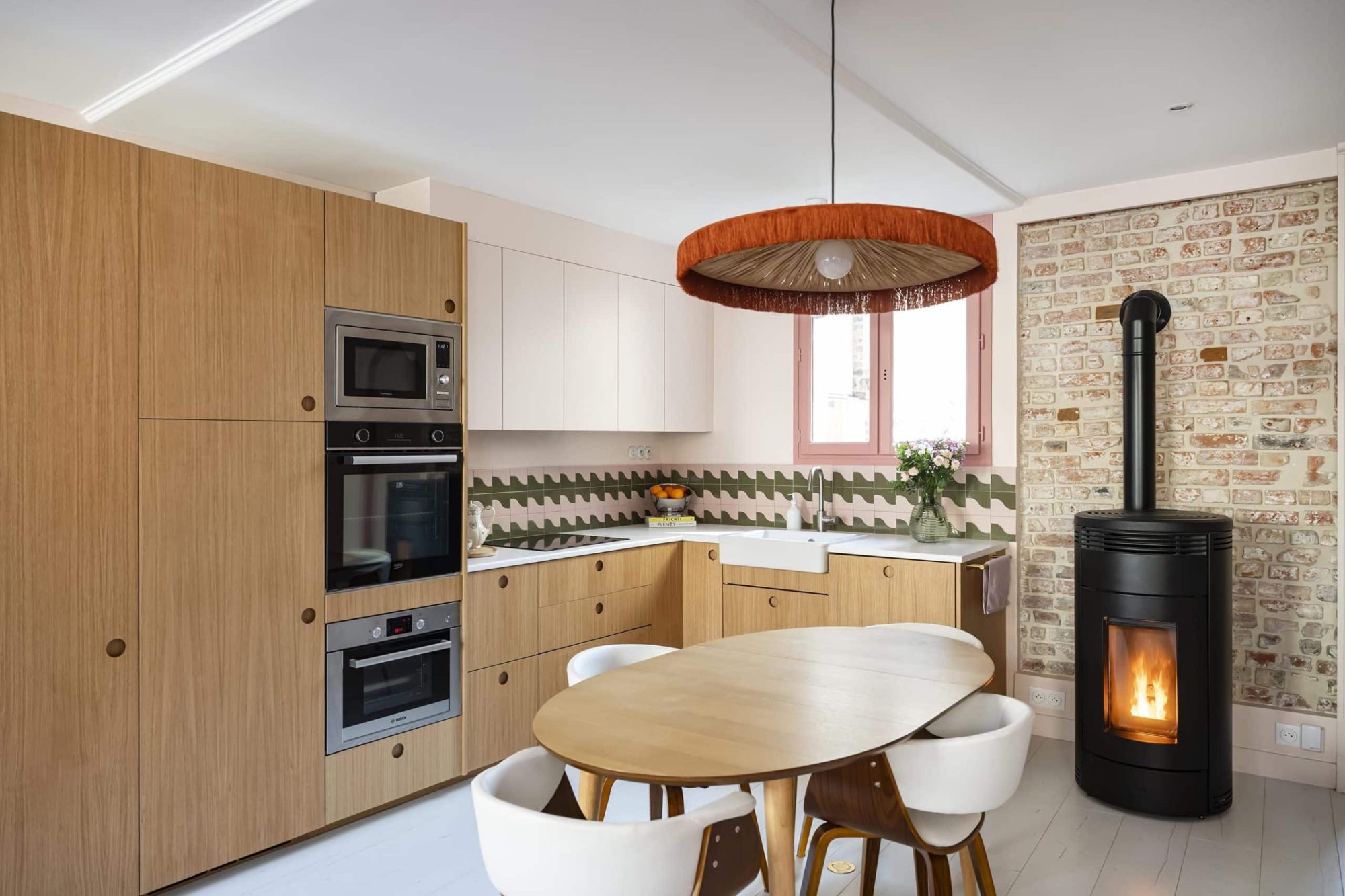 Cuisine ouverte avec façades en bois, table ronde, suspension et poêle noir contre un mur en briques, rénovation d'une maison à Colombes par Nathalie Bertaux, architecte d'intérieur